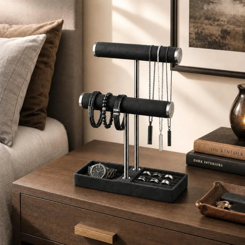 Black 2‑tier jewelry display stand on a bedside dresser holding necklaces, bracelets, rings, and a watch in a styled bedroom scene.