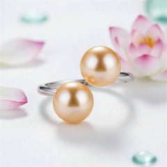 Sterling silver ring featuring two gold pearls resting on a glossy pearl‑white surface, with soft magenta, blush, and aqua accents and a white lotus flower with pink‑magenta–tipped petals softly blurred in the background.