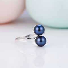 Sterling silver ring featuring two deep midnight‑blue pearls resting on a glossy white marble surface, with soft peach and mint rounded shapes blurred in the background.