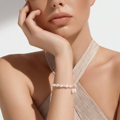 Close-up of a woman wearing a white freshwater pearl bracelet with two dangling pearls, shown in soft lighting against a neutral background while she rests her hand gently near her face.