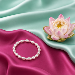 White pearl bracelet displayed beside a pink lotus flower on mint‑aqua and lotus‑magenta satin fabric, highlighting the pearls’ luster and brand elegance.