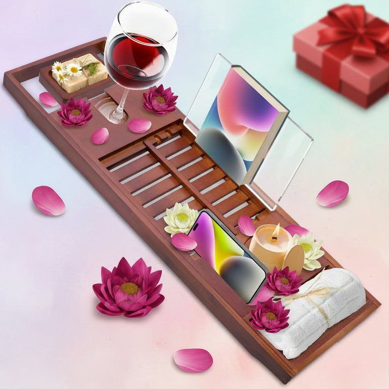 Wooden bathtub caddy tray with wine glass, soap, flowers, lit candle, rolled towel, smartphone, and book arranged for a luxurious spa-style bath setup